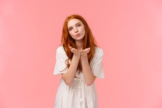 Waist-up portrait, cute and sensual, romantic feminine girl with red curly hair in white dress, bending and holding palms near folded lips, sending air kiss at camera, standing pink background.