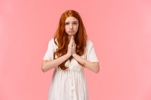 Gloomy and sad, cute pouting redhead girl asking for help, looking hopeful camera, make promise or appologizing, press hands together in pray, begging favour, pleading over pink background.