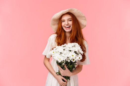 Excited joyful charismatic redhead woman having perfect date, feeling happy and carefree, holding bouquet white flowers and laughing cheerful, wearing sun hat, standing pink background.