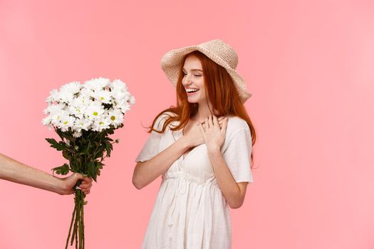 Gifts, celebration and tenderness concept. Surprised cute, alluring redhead woman in hat, dress, charmed with pleasant gift, holding hands on heart flattered, smiling, looking at bouquet flowers.