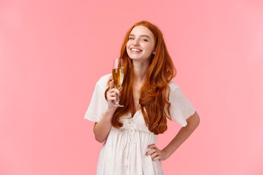 Happiness, confident and celebration concept. Happy and carefree alluring redhead b-day girl sipping champagne and laughing, having fun, enjoying party, standing over pink background.