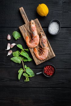Jumbo shrimps on wooden board with herbs over black wood table, top view, flat lay, food photo.
