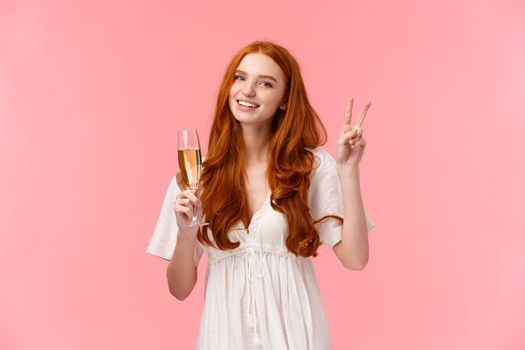 Lovely romantic caucasian redhead female in white cute dress, showing peace sign, informal greeting of friend on party, smiling holding glass of champagne, drinking as celebrating, attend event.