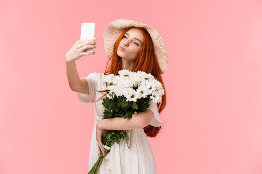 Silly and cute, feminine girl blogger taking selfie with bouquet flowers boyfriend brought, holding smartphone folding lips in air kiss, posing for perfect sensual shot, stand pink background.