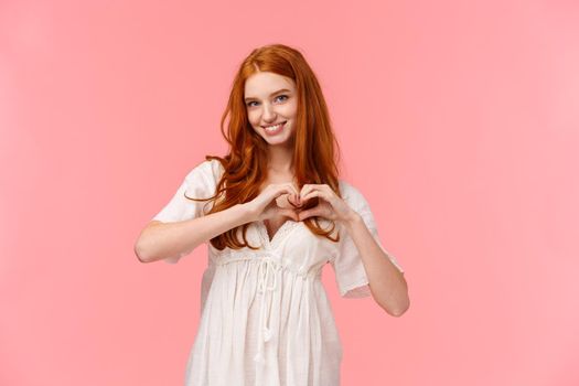 Waist-up portrait lovely, sensual redhead girlfriend in white dress, showing her love and care, make heart gesture and smiling, confess affection, sincere sympathy, pink background.