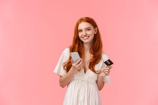 Shopping, internet and mobile concept. Attractive lovely redhead female in white dress, holding smartphone and credit card, purchase online, buying something or sending money to friend.