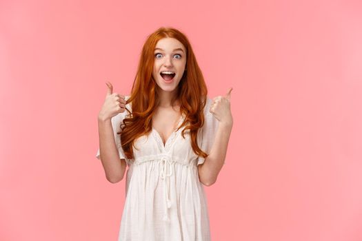 Discounts, shopping and people concept. Cheerful redhead woman amazed with wonderful event, attend awesome concert, show thumbs-up and smiling astonished, pink background.