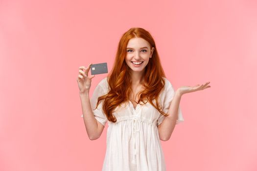 Payment and purchases now so simple. Easy banking system concept. Attractive pleasant caucasian redhead girl introduce credit card and smiling satisfied, telling about bank pros, pink background.