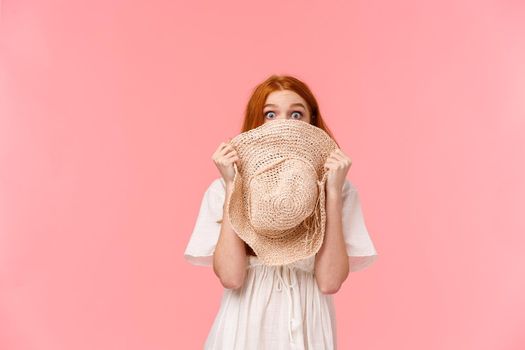Surprise, look at camera. Shy and blushing cute startled redhead girl hiding face behind straw hat, staring camera scared or shook, standing pink background overwhelmed.