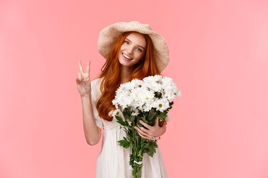 Spring, celebration and emotions concept. Cheerful lovely redhead female in cute straw hat, enjoying romantic date, holding bouquet white flowers, show peace sign and smiling happy, pink background.