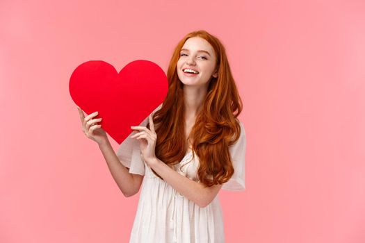 Happiness, love and relationship concept. Lovely and cute redhead teenage girl laughing and smiling camera, holding big red heart joyfully expressing sympathy, deep feelings, pink background.