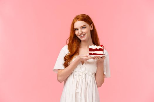 Lovely, romantic and alluring redhead girlfriend baked delicious surprise for valentines day date, holding peace cake on plate and smiling tempting with sassy, coquettish expression, pink background.