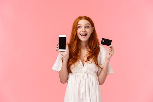 Alluring, delighted and fascinated cute happy redhead girl smiling satisfied and amused, holding credit card, showing her smartphone display, made great purchase, bragging to friend, pink background.