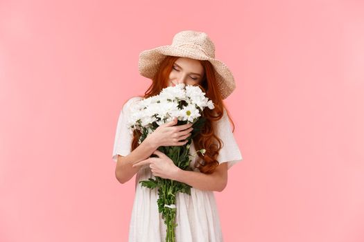 Romance, love and relationship concept. Romantic dreamy, alluring redhead girl sniffing flowers, close eyes and hugging beautiful bouquet, standing pink background. Copy space