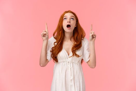Wow so cool, check it out. Waist-up portrait speechless, curious and fascinated redhead caucasian female in white dress, looking pointing up, showing top advertisement, thrilled over pink background.