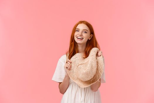 Waist-up shot tender, charismatic and alluring redhead girl packed-up for summer vacation, ready to travel abroad, holding straw hat, laughing and smiling camera. standing pink background.