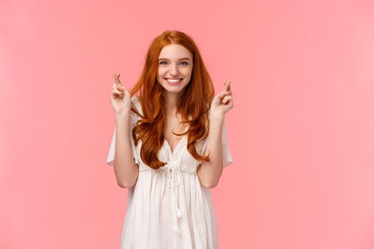 Confident, optimistic and hopeful cute redhead female in white dress, cross fingers good luck and smiling, making wish, anticipate dream come true, standing pink background upbeat.