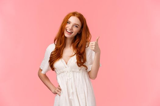 Confident good-looking redhead optimistic girl, saying everything be alright, all good, showing thumb-up and smiling, nod in approval, agree on terms, recommend product, pink background.