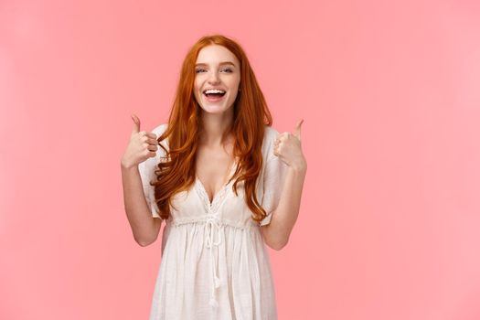 Waist-up portrait cheerful, upbeat redhead woman in white dress, showing thumbs-up and smiling in approval, like product, recommend it, express positive opinion, standing pink background.