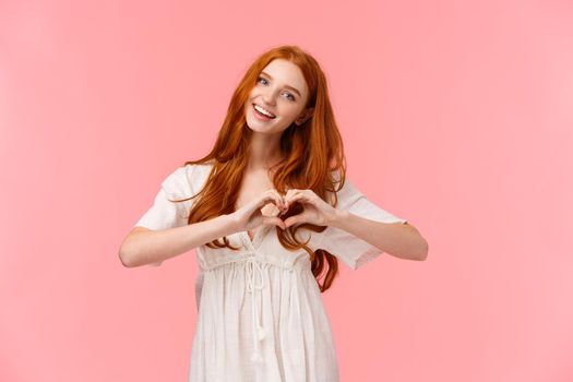 Waist-up portrait lovely, romantic and cute european woman with red hair, tilt head and smiling, saying love you, make heart gesture in like, express affection or sympathy, happy valentines day.