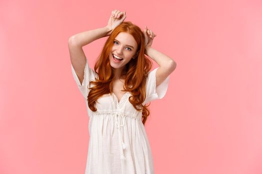 Charming, pretty and cute redhead female mimicking dog, holding hands over head as puppy ears, tilt head and smiling, playfully fool around, feeling goofy, standing pink background kawaii pose.