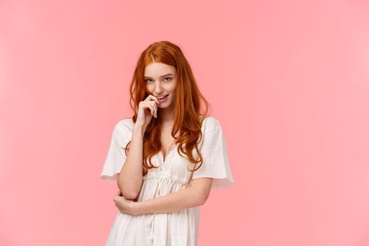 Sensual, romantic and sassy, seductive redhead daring woman in white dress, biting finger and coquettish look camera from under forehead, standing pink background, flirting.