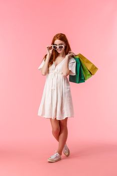 Full-length vertical shot alluring happy redhead female release stress with shopping, shopaholic walking along stores, shop-malls with bags behind back, take-off glasses, smiling camera.