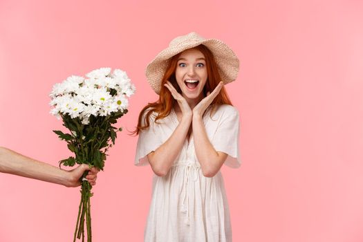 Surprises, celebration and holidays concept. Astonished happy and cheerful smiling redhead girl scream from happiness and joy, looking enthusiastic as someone giving her flowers, pink background.