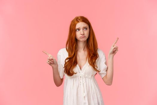 Waist-up portrait innocent, cute and timid redhead indecisive girl, dont know what do, pouting feeling sad, gloomy look around, pointing fingers left and right, pink background.