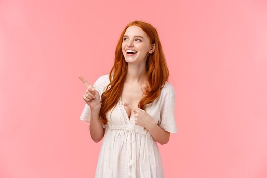 Charismatic lovely european redhead woman in white trendy dress, showing something hilarious, pointing and looking upper left corner, laughing out loud, express happiness and joy.