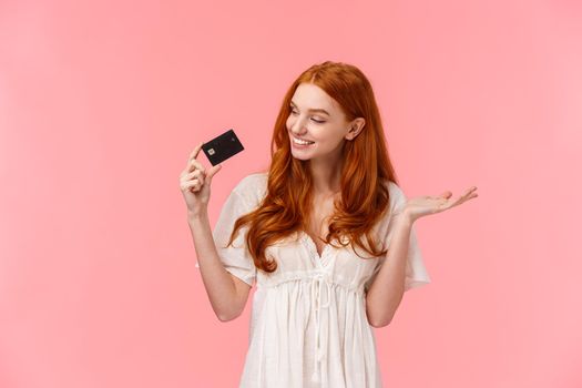 Satisfied cute redhead woman praise her credit card, looking at it pleased and happy, smiling, wasting money on bank account, receive payment and going shopping, standing pink background.
