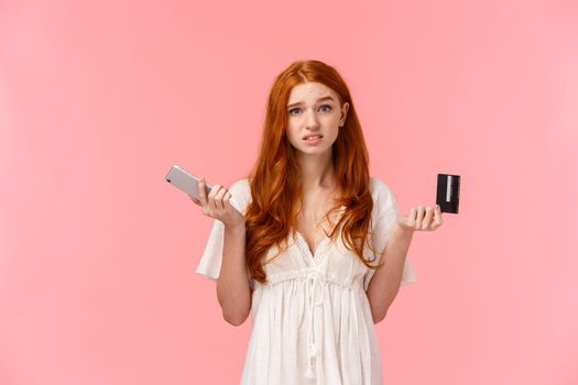 Indecisive and unsure cute european redhead woman shrugging, looking confused and useless camera, shrugging with smartphone and credit card in hands, dont know what order online.