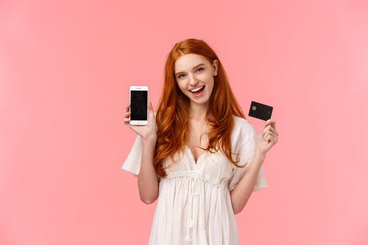 Excited, sassy, carefree female with red curly hair like wasting money in internet store, making order, got tickets in flight app, holding credit card and smartphone, showing promo on mobile screen.