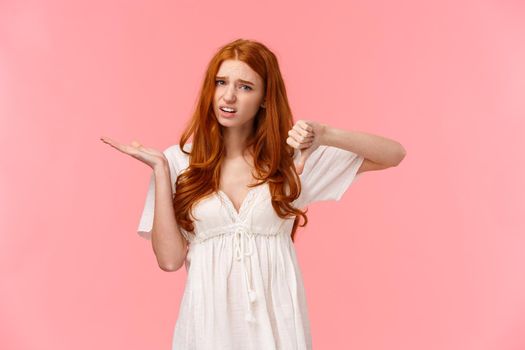 Skeptical displeased redhead female customer have her negative opinion, pointing at something bothering and really bad, grimacing unsatisfied, show thumb-down in dislike, disagree gesture.