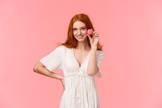 Beauty, desserts and people concept. Alluring and sassy redhead caucasian girl in white dress, seducing someone with tasty, delicious macaron, smiling inviting try, pink background.