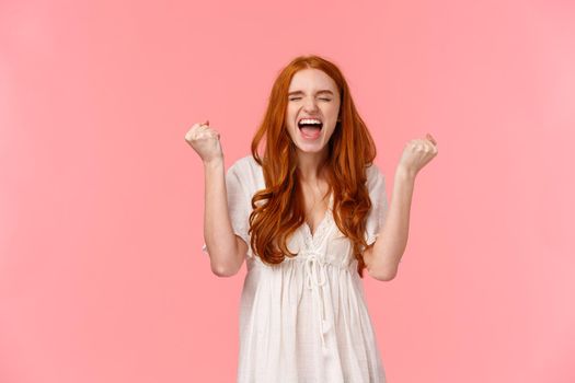 Challenge, celebration and victory concept. Cheerful delighted lucky redhead girl, feeling extremely happy winning prize, fist pump triumphing, yelling hooray or yes, standing pink background.