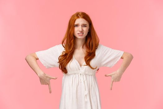 Well I dont know. Waist-up portrait indecisive and unsure, slightly displeased cute redhead woman in white dress, pointing down, cringe as express dislike, disappointed with gift, pink background.