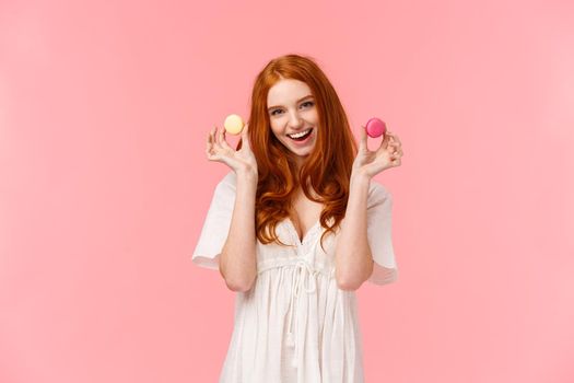 Girl inviting sit with her enjoy tea with desserts. Stunning, alluring redhead female in white dress, showing two macarons tempting friend have bite, smiling excited, standing pink background.