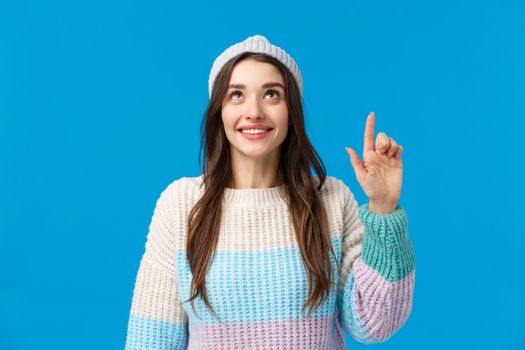 Dreamy beautiful young brunette woman in winter sweater, hat, smiling, looking and pointing up with happy cheerful expression, contemplate something mesmerizing, found excellent choice.