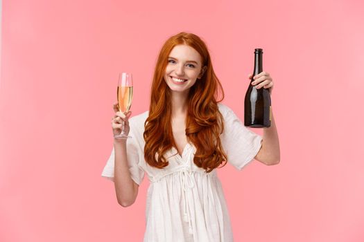 Cheers, congratulations my friend. Tender and happy, friendly-looking charming redhead woman in white dress, raising glass and holding champagne bottle, celebrating success, pink background.