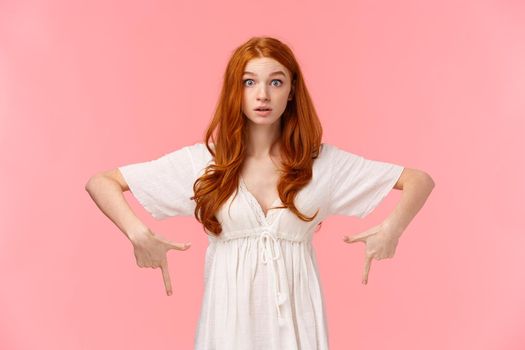 Curious and intrigued redhead girl heard interesting news, raise eyebrows in wonder and amazement, pointing fingers down as asking question about product, stand pink background.