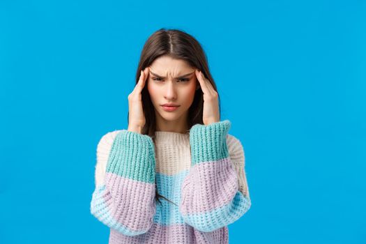 Waist-up portrait tensed young woman feeling unwell, touching temples and squinting, having huge hungover, painful feelings in head, suffering migraine, headache standing blue background.