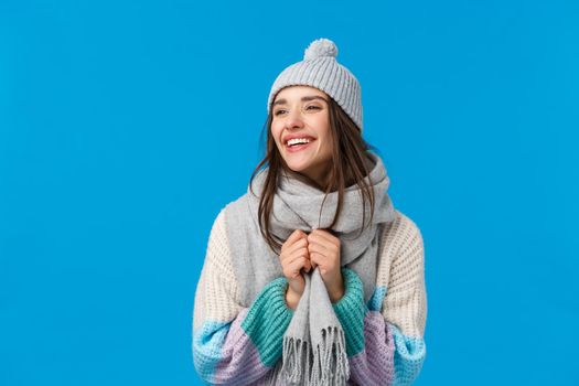 Carefree cheerful laughing and smiling brunette woman in winter hat, scarf and sweater, enjoying christmas holidays, playing outside in snow, standing joyful over blue background.