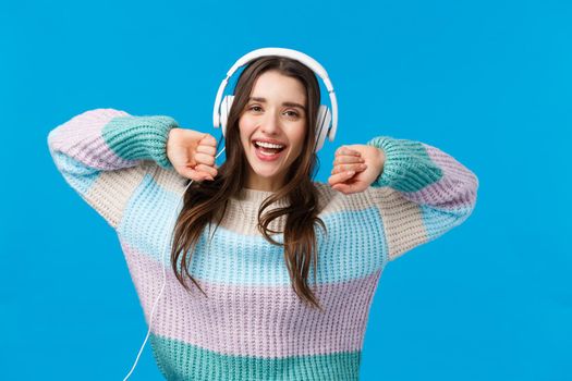 Waist-up portrait carefree joyful girl home alone, enjoying freedom and awesome music in good quality headphones, dancing raising hands up cheerful, smiling camera, blue background.