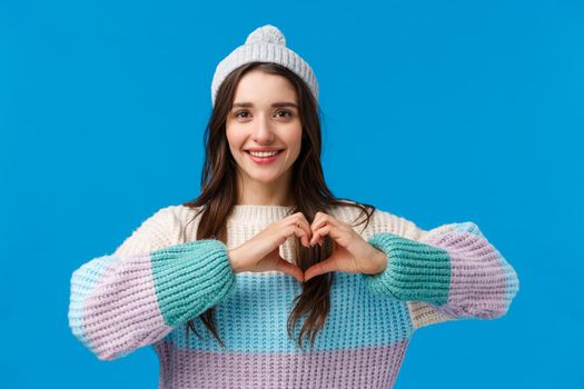 Valentines day, relationship and emotions concept. Lovely, pretty brunette girl in winter hat, sweater, showing heart sign and smiling, looking camera happy, show love and affection, blue background.