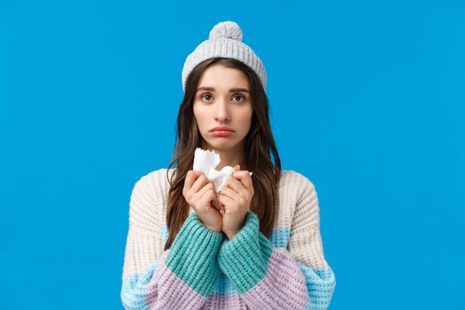 Upset young woman feelings unwell, have fever, sick on winter holidays, wearing hat and sweater, holding napkin sneeze and have runny nose, looking sad camera, caught cold or flu, blue background.