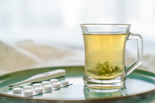 Glass transparent cup with herbal tea and grass leaves, thermometer, tablets on a green tray. Concept of health