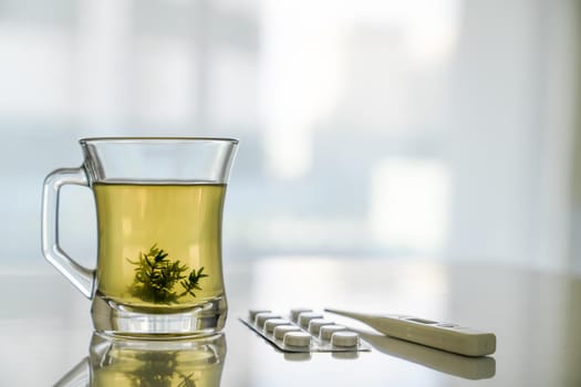 Glass transparent cup with herbal tea and grass leaves, thermometer, tablets on white table. Concept of health treatment