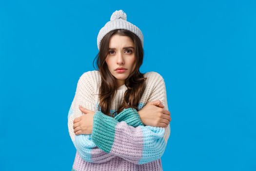 Turn on heater please its freezing. Upset and gloomy young silly girl in winter hat, sweater, trembling from low temprature, hugging herself warm-up, hate cold season, blue background.