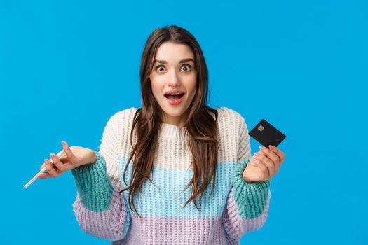 Surprised and wondered, amused cute caucasian woman in winter sweater, shrugging amazed, holding credit card and smartphone, got cashback, standing blue background joyful.